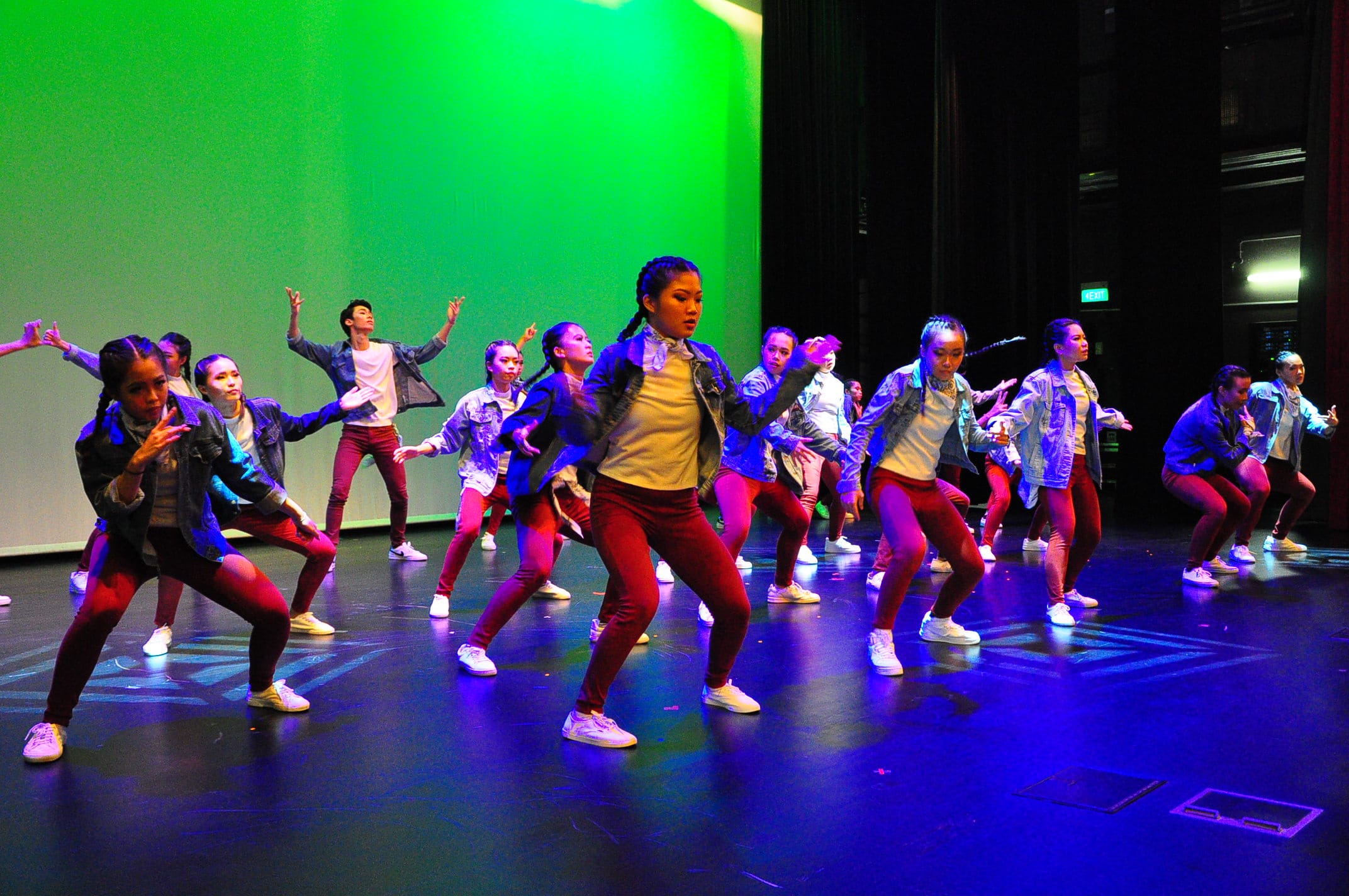 A large group of dancers in denim jackets and red pants perform on a stage lit with green and blue lights.